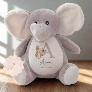 Peut inclure: Une peluche &eacute;l&eacute;phant grise avec de grandes oreilles et un ventre blanc. Le ventre pr&eacute;sente un motif floral avec un lapin, la lettre "A", le nom "Annie" et la date "16.04.2025". Un autocollant rose avec "Indie Prints" est &agrave; gauche.