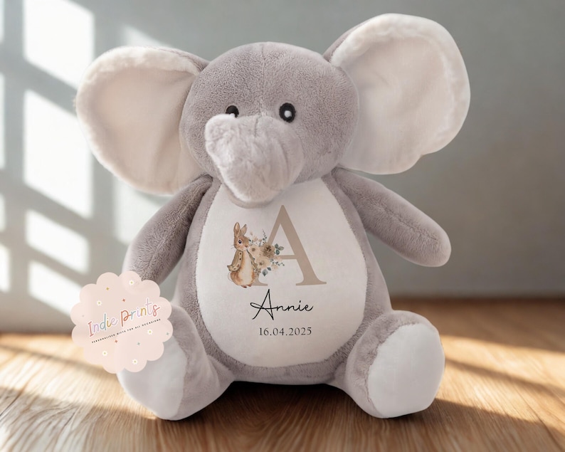 Peut inclure: Une peluche &eacute;l&eacute;phant grise avec de grandes oreilles et un ventre blanc. Le ventre pr&eacute;sente un motif floral avec un lapin, la lettre "A", le nom "Annie" et la date "16.04.2025". Un autocollant rose avec "Indie Prints" est &agrave; gauche.
