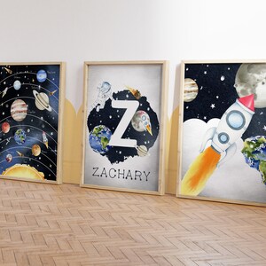 May include: Three framed watercolor art prints with a space theme. One shows the solar system, another features the letter "Z" with planets and an astronaut, and the third depicts a rocket ship. The prints are in wooden frames.
