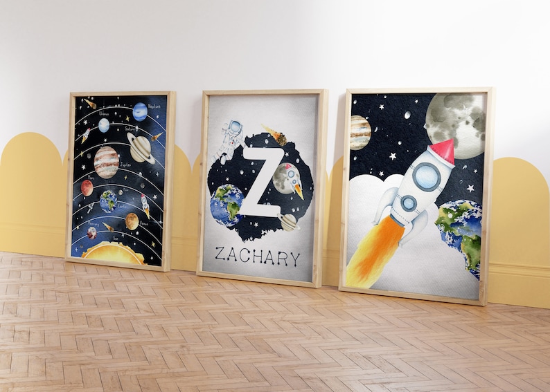 May include: Three framed watercolor art prints with a space theme. One shows the solar system, another features the letter "Z" with planets and an astronaut, and the third depicts a rocket ship. The prints are in wooden frames.