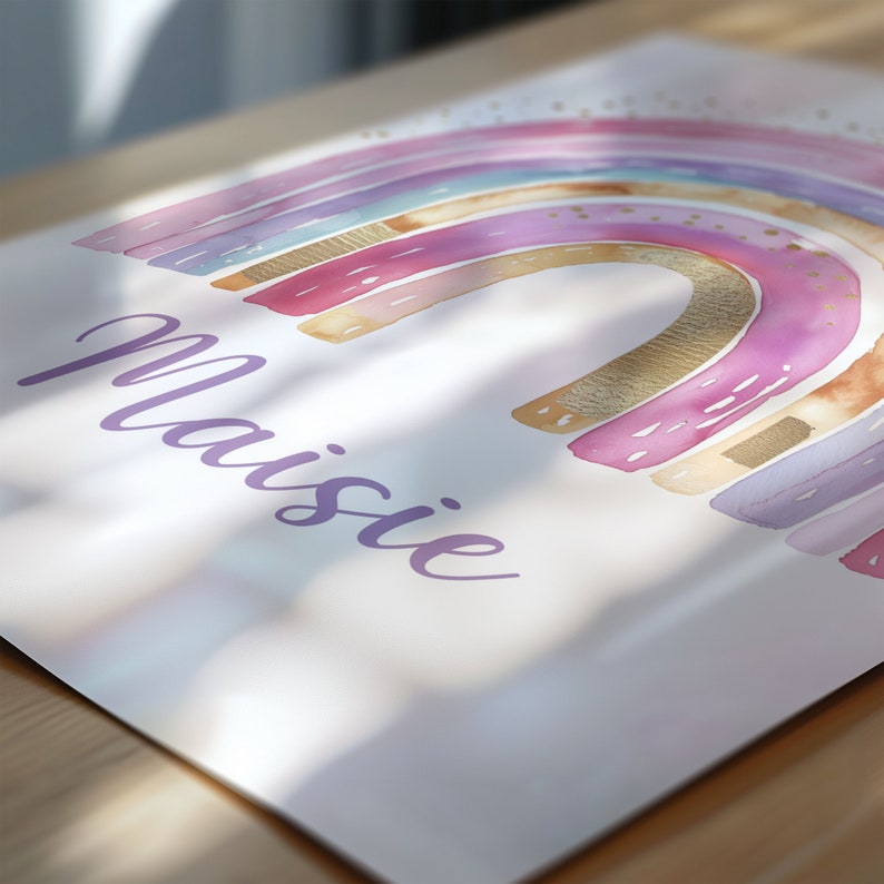 Puede incluir: Una impresi&oacute;n de acuarela con un dise&ntilde;o de arco&iacute;ris en tonos rosa, morado y dorado. El nombre "Maisie" est&aacute; escrito en cursiva debajo del arco&iacute;ris. La obra de arte est&aacute; sobre un fondo blanco.