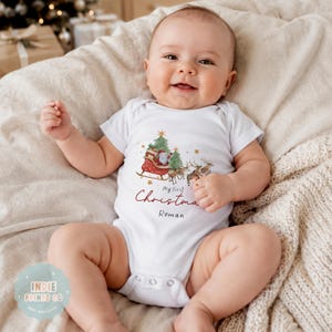 May include: A white short-sleeved baby bodysuit featuring a watercolor illustration of Santa Claus in a sleigh pulled by reindeer, with the text "My First Christmas" in red script and the name "Roman" printed in black below the graphic.