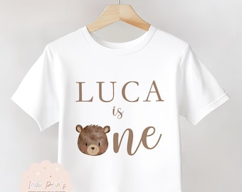 May include: White t-shirt with the name "LUCA" in brown letters, the word "is", and the word "one". A brown bear graphic is below the name. The shirt is hanging on a wooden hanger.