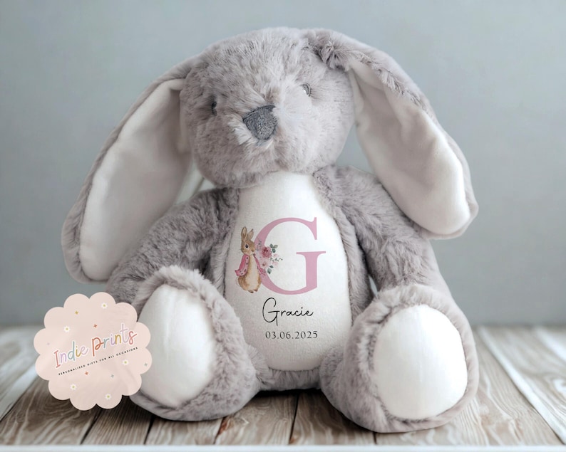 Peut inclure: Une peluche lapin grise avec de longues oreilles et l'int&eacute;rieur blanc. Le ventre du lapin est blanc avec un motif floral rose, la lettre "G" et le nom "Gracie" avec une date. Un jouet en peluche.