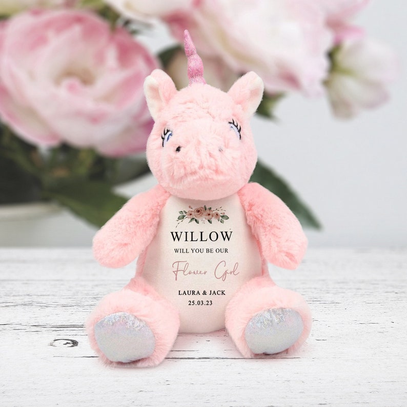 Pode incluir: Um brinquedo de unic&oacute;rnio de pel&uacute;cia rosa com um chifre rosa, c&iacute;lios azuis e cascos prateados. O unic&oacute;rnio tem uma barriga branca com o texto "WILLOW, WILL YOU BE OUR Flower Girl?" e os nomes "LAURA & JACK" com a data "25.03.23".