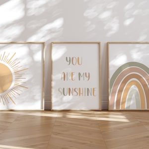 May include: Three framed art prints with a sun, a rainbow, and the text "YOU ARE MY SUNSHINE". The prints have a neutral color palette with beige, orange, and green hues. The frames are a light wood color.