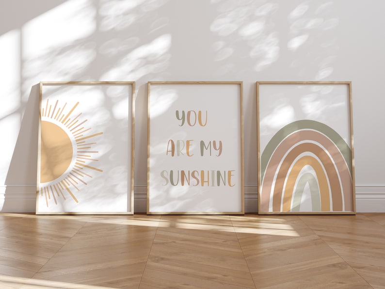 May include: Three framed art prints with a sun, a rainbow, and the text "YOU ARE MY SUNSHINE". The prints have a neutral color palette with beige, orange, and green hues. The frames are a light wood color.