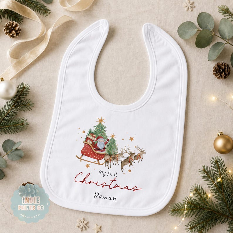 May include: A white baby bib featuring a watercolor illustration of Santa Claus in a red sleigh pulled by reindeer, with two green Christmas trees. The text reads "My First Christmas" in red script and the name "Roman" in black lettering below.