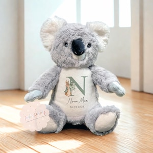 Peut inclure: Une peluche koala grise avec un ventre blanc orn&eacute; d'un motif floral, de la lettre "N" et du nom "Nova Mae" avec la date "26.05.2025". Le koala a un nez noir et des oreilles blanches. Un autocollant avec le texte "Indie Prints" est visible.