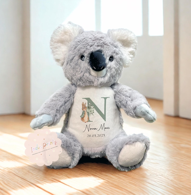 Peut inclure: Une peluche koala grise avec un ventre blanc orn&eacute; d'un motif floral, de la lettre "N" et du nom "Nova Mae" avec la date "26.05.2025". Le koala a un nez noir et des oreilles blanches. Un autocollant avec le texte "Indie Prints" est visible.