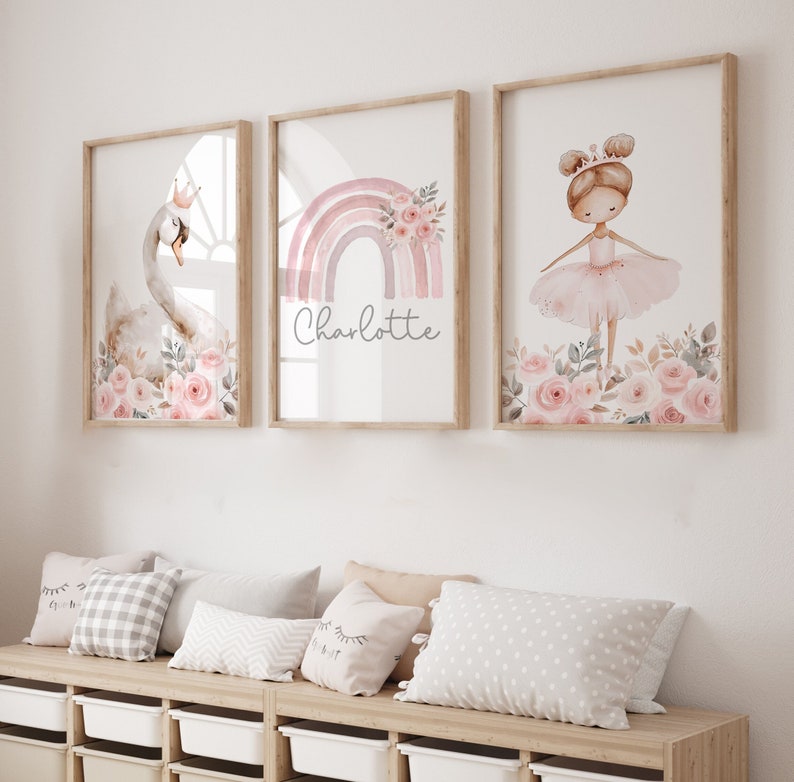 May include: Three framed art prints with a swan, a rainbow with the name "Charlotte", and a ballerina. The prints feature soft pink and beige tones, floral accents, and are displayed above a bench with pillows.