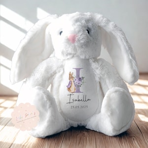 Peut inclure: Une peluche blanche en forme de lapin avec de longues oreilles et un nez rose. Le lapin porte une personnalisation sur le ventre avec le nom "Isabella", la date "25.05.2025" et un motif floral et lapin.