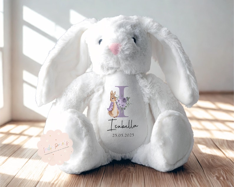 Peut inclure: Une peluche blanche en forme de lapin avec de longues oreilles et un nez rose. Le lapin porte une personnalisation sur le ventre avec le nom "Isabella", la date "25.05.2025" et un motif floral et lapin.