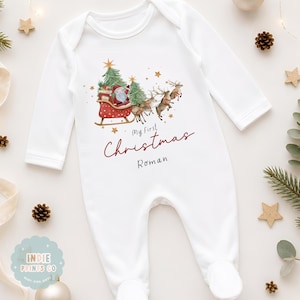 May include: A white long-sleeve baby bodysuit featuring a watercolor illustration of Santa Claus in a red sleigh pulled by reindeer among green pine trees and gold stars. The text reads My First Christmas and the name Roman in black script.