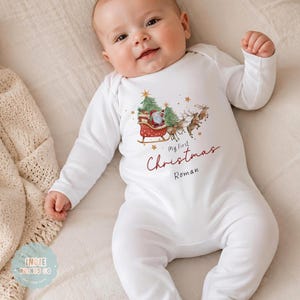 May include: A white long-sleeved baby bodysuit featuring a graphic of Santa Claus in a sleigh pulled by reindeer, two pine trees, and gold stars. The text reads My First Christmas and the name Roman in a dark red and black script font.