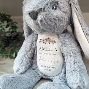 Pode incluir: Um coelho de pel&uacute;cia cinzento com orelhas compridas e uma barriga branca. A barriga apresenta o texto "AMELIA WILL YOU BE OUR Flower Girl" com os nomes Jessica & Paul e a data 17.09.23. Acentos florais adornam o texto.