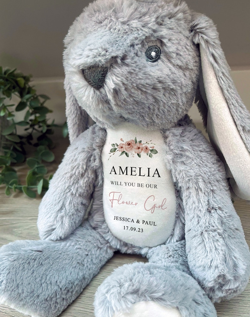 Pode incluir: Um coelho de pel&uacute;cia cinzento com orelhas compridas e uma barriga branca. A barriga apresenta o texto "AMELIA WILL YOU BE OUR Flower Girl" com os nomes Jessica & Paul e a data 17.09.23. Acentos florais adornam o texto.