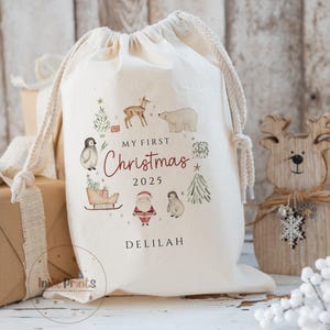 May include: A cream-coloured drawstring Christmas gift bag featuring festive illustrations of animals and trees. The text "MY FIRST Christmas 2025" and a name are printed on the front. The bag is made of a soft fabric and has a drawstring closure.