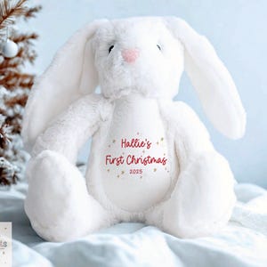 May include: A white plush bunny with long ears and a pink nose. The bunny has the text "Hallie's First Christmas 2025" printed on its chest. The bunny is sitting on a white blanket with a Christmas tree in the background.