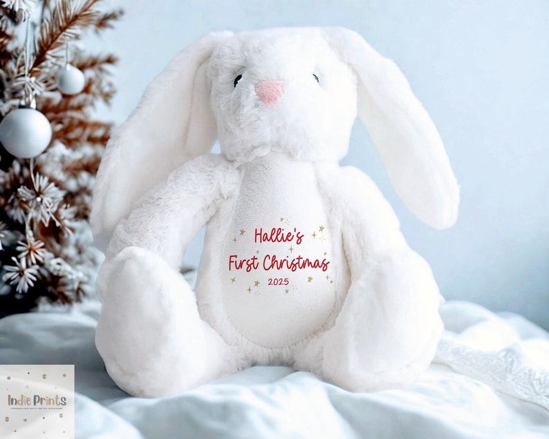 May include: A white plush bunny with long ears and a pink nose. The bunny has the text "Hallie's First Christmas 2025" printed on its chest. The bunny is sitting on a white blanket with a Christmas tree in the background.