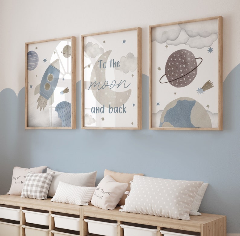 May include: Three framed art prints with a space theme. The prints feature a rocket, a crescent moon with the text "To the moon and back", and planets. The color palette includes blue, gray, and beige tones. The frames are light brown.