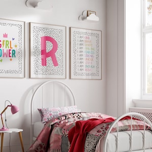 May include: Three framed art prints with white backgrounds and black polka dots. One print says "GIRL POWER" in colorful letters. Another features a large pink letter "R". The third print lists positive affirmations. A white bed frame and bedding are in the foreground.