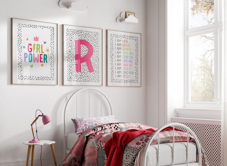 May include: Three framed art prints with white backgrounds and black polka dots. One print says "GIRL POWER" in colorful letters. Another features a large pink letter "R". The third print lists positive affirmations. A white bed frame and bedding are in the foreground.