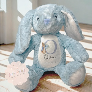 Peut inclure: Un lapin en peluche bleu clair avec de longues oreilles. Le lapin a un ventre blanc avec un motif personnalis&eacute; avec un "C" floral, une illustration de Pierre Lapin, le nom "Oliver" et la date "22.03.2025". Le lapin est pos&eacute; sur une surface en bois.