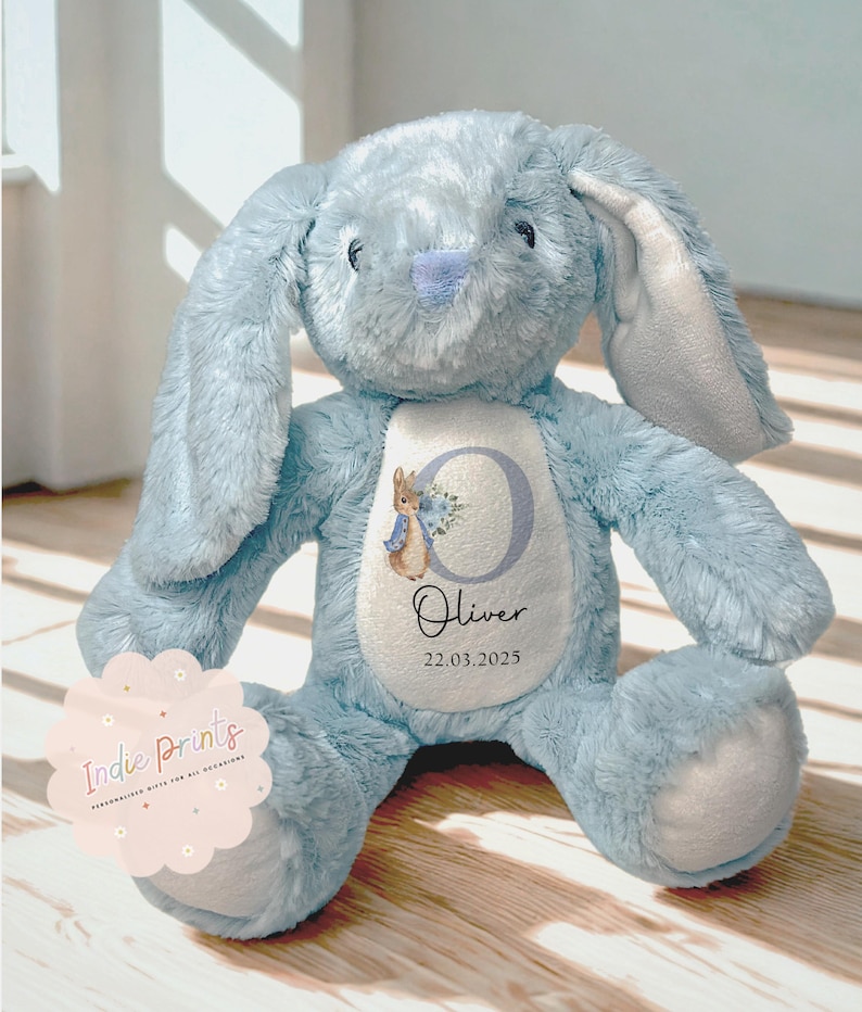 Peut inclure: Un lapin en peluche bleu clair avec de longues oreilles. Le lapin a un ventre blanc avec un motif personnalis&eacute; avec un "C" floral, une illustration de Pierre Lapin, le nom "Oliver" et la date "22.03.2025". Le lapin est pos&eacute; sur une surface en bois.