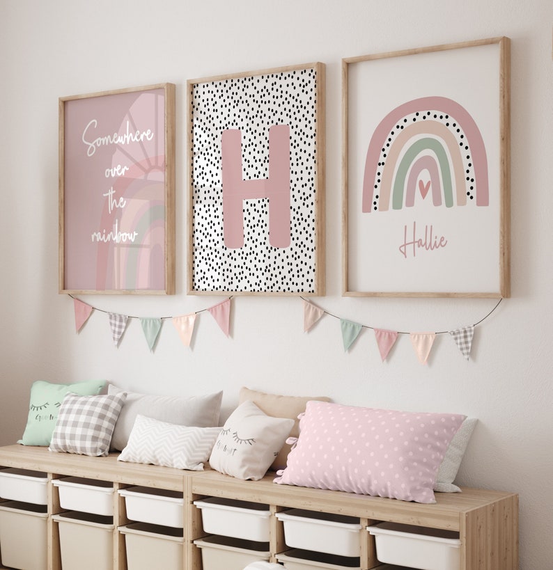 May include: Three framed wall art prints with a rainbow, an initial, and the text "Somewhere over the rainbow." The prints are in soft pink and neutral tones. A garland of pastel-colored pennants hangs below, with pillows and storage bins below.