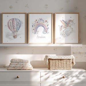 Op de afbeelding: Drie ingelijste aquarelprints met een luchtballon, een regenboog met de naam "Amelia" en een eenhoorn. De prints worden tentoongesteld op een witte plank boven een witte dressoir. Een stapel opgevouwen dekens en een rieten mand staan op de dressoir.