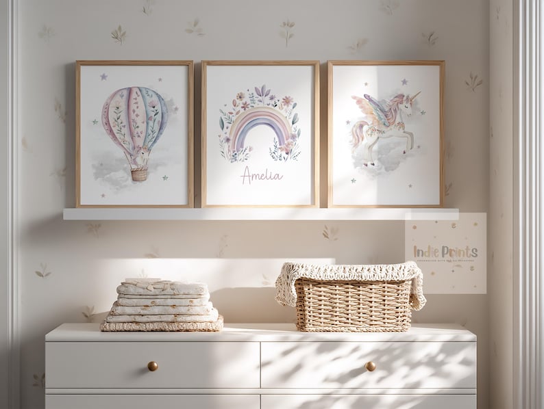 Op de afbeelding: Drie ingelijste aquarelprints met een luchtballon, een regenboog met de naam "Amelia" en een eenhoorn. De prints worden tentoongesteld op een witte plank boven een witte dressoir. Een stapel opgevouwen dekens en een rieten mand staan op de dressoir.