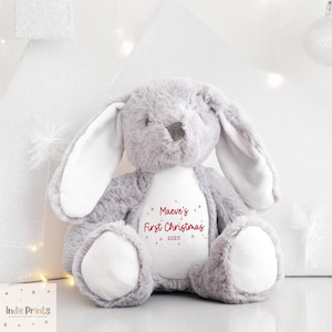 May include: A gray plush bunny rabbit with long ears and white inner ears. The bunny has a white belly with the text "Maeve's First Christmas 2025" in red script. The bunny is a Christmas-themed plush toy.