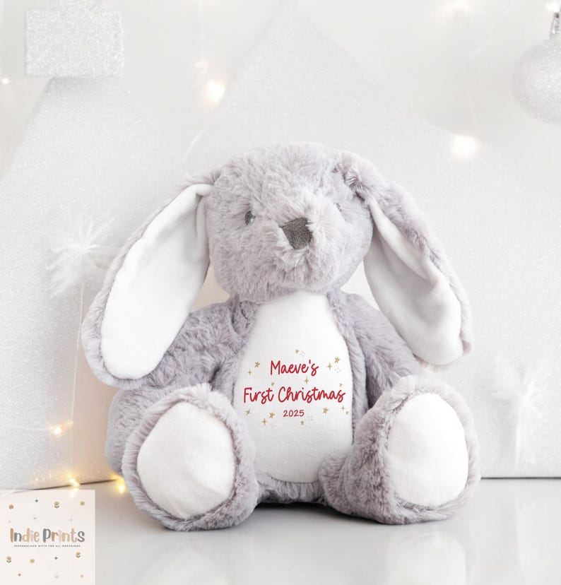 May include: A gray plush bunny rabbit with long ears and white inner ears. The bunny has a white belly with the text "Maeve's First Christmas 2025" in red script. The bunny is a Christmas-themed plush toy.