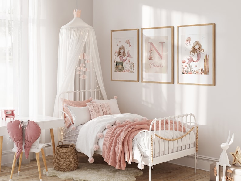 May include: A child's bedroom with a white metal bed, adorned with pink and white bedding and a matching throw blanket. Three framed mermaid art prints hang above the bed. A white canopy with a crown and hanging decorations adds a whimsical touch.