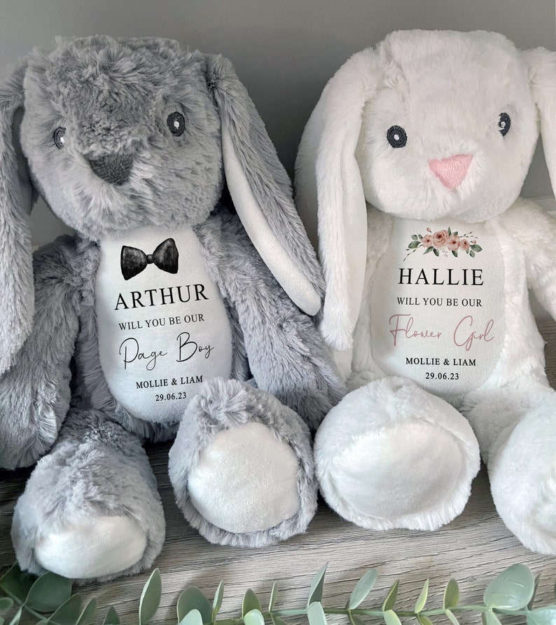 Pode incluir: Dois coelhos de pel&uacute;cia, um cinza e outro branco, com texto personalizado. O coelho cinza tem uma gravata borboleta preta e diz "ARTHUR WILL YOU BE OUR Page Boy 29.06.23". O coelho branco diz "HALLIE WILL YOU BE OUR Flower Girl 29.06.23".