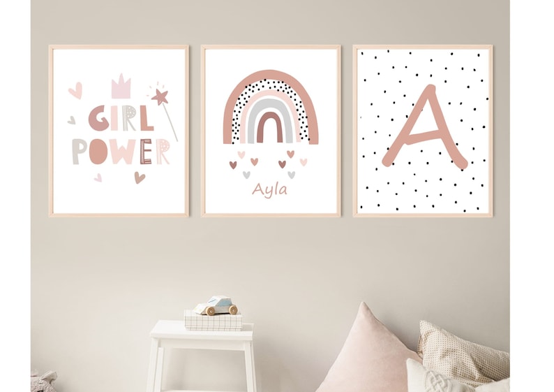 May include: Three framed art prints with a nursery theme. The prints feature a pink color palette with the words "GIRL POWER", a rainbow with the name "Ayla", and the letter "A" with polka dots. The frames are light wood.