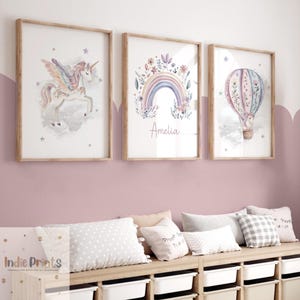 Op de afbeelding: Drie ingelijste aquarelprints. De prints tonen een eenhoorn, een regenboog met de naam "Amelia" en een heteluchtballon. Het kunstwerk is in zachte pastelkleuren, met houten frames. De prints worden weergegeven boven een houten plank met kussens en opbergbakken.