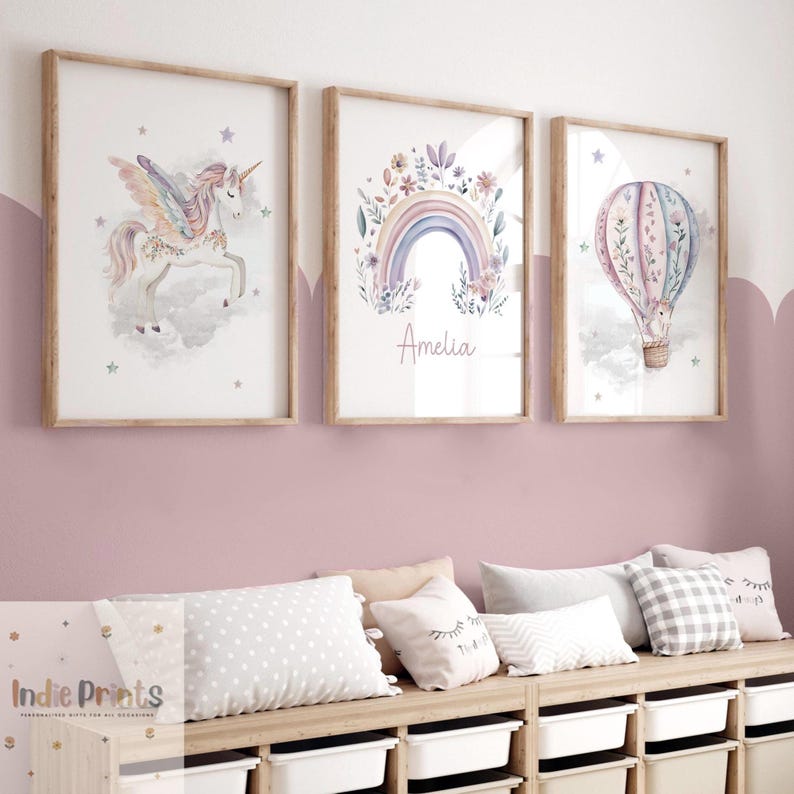 Op de afbeelding: Drie ingelijste aquarelprints. De prints tonen een eenhoorn, een regenboog met de naam "Amelia" en een heteluchtballon. Het kunstwerk is in zachte pastelkleuren, met houten frames. De prints worden weergegeven boven een houten plank met kussens en opbergbakken.