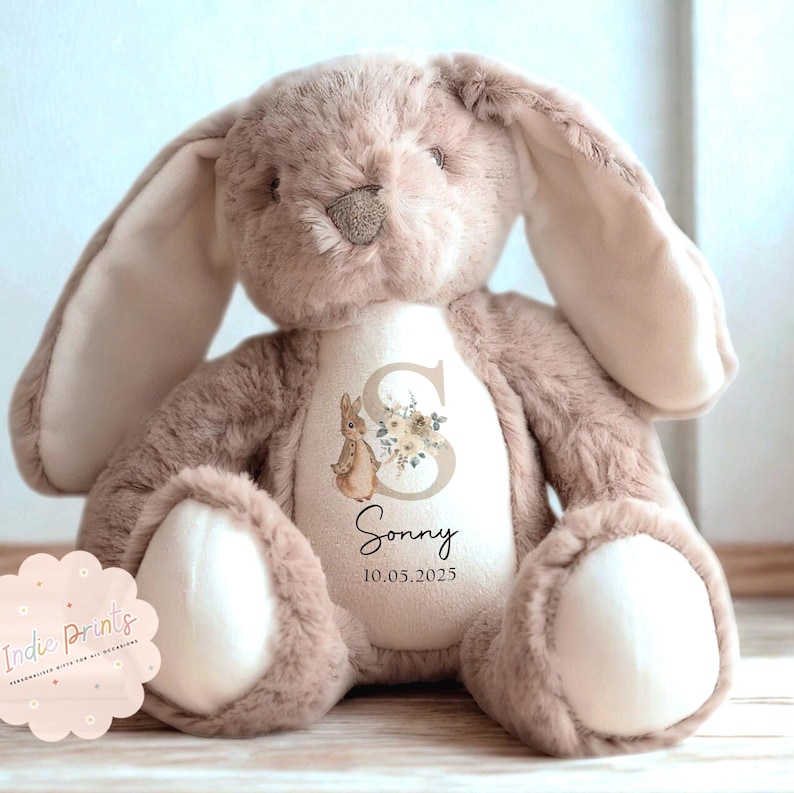 Peut inclure: Une peluche en forme de lapin dans les tons taupe et cr&egrave;me. Le lapin a de longues oreilles tombantes et un aspect doux et c&acirc;lin. La poitrine du lapin pr&eacute;sente un motif floral avec la lettre "S", une illustration de lapin, le nom "Sonny" et la date "10.05.2025".