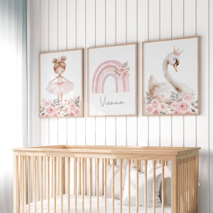 May include: Three framed art prints with a nursery theme. The prints feature a ballerina, a rainbow with the name "Vienna", and a swan wearing a crown, all in soft pink and beige tones. The artwork is displayed above a wooden crib.