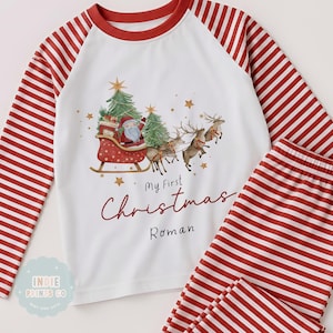 May include: A white long-sleeve pajama set with red and white striped sleeves and pants. The shirt features a graphic of Santa in a sleigh pulled by reindeer, green Christmas trees, and gold stars. Text reads My First Christmas Roman.