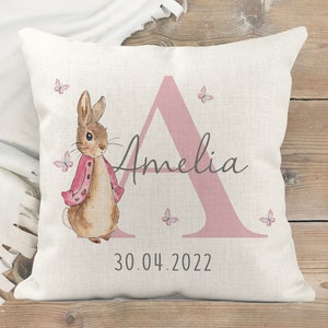 Puede incluir: Un cojín cuadrado de color crema con una ilustración de acuarela de un conejo con una chaqueta rosa, con el nombre "Amelia" y la fecha "30.04.2022". Mariposas rosas y una gran letra "A" rosa completan el diseño.