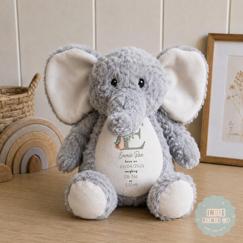 May include: A plush gray elephant toy with large cream-colored inner ears and a white belly patch. The belly features the letter E, a pear illustration, and the text: Emmie Rae, born on 03/04/2026, weighing 5lb 7oz at 3.12am.