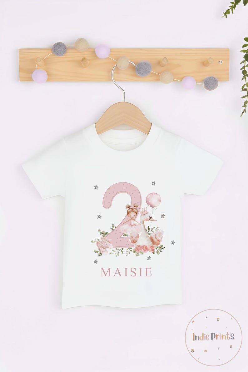 Pu&ograve; includere: T-shirt bianca con un disegno rosa del numero 2 con una principessa, un cigno ed elementi floreali. Il nome "Maisie" &egrave; stampato sotto il disegno. La maglietta &egrave; appesa a un appendiabiti di legno.