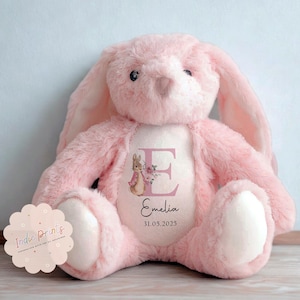 Peut inclure: Une peluche rose en forme de lapin avec de longues oreilles et un ventre blanc. Le lapin porte une personnalisation sur sa poitrine avec la lettre "E", une illustration florale, le nom "Emelia" et la date "31.05.2025".
