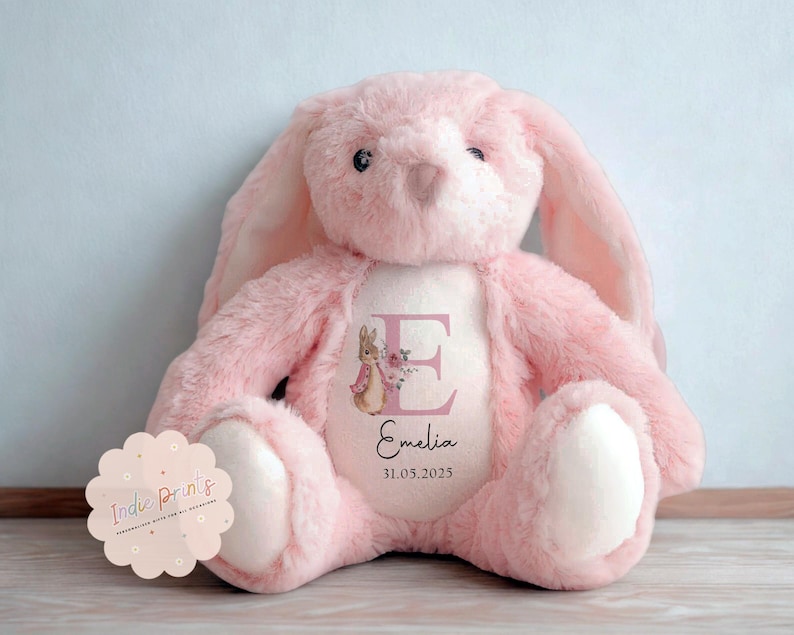 Peut inclure: Une peluche rose en forme de lapin avec de longues oreilles et un ventre blanc. Le lapin porte une personnalisation sur sa poitrine avec la lettre "E", une illustration florale, le nom "Emelia" et la date "31.05.2025".