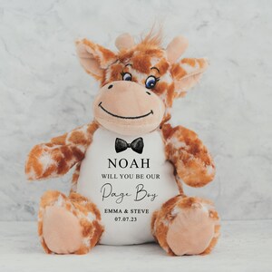 Pode incluir: Um brinquedo de pel&uacute;cia de girafa com manchas marrons e brancas, uma gravata borboleta preta e o texto "NOAH WILL YOU BE OUR Page Boy" com nomes e uma data. A girafa tem um rosto sorridente e est&aacute; sentada.