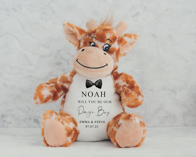 Pode incluir: Um brinquedo de pel&uacute;cia de girafa com manchas marrons e brancas, uma gravata borboleta preta e o texto "NOAH WILL YOU BE OUR Page Boy" com nomes e uma data. A girafa tem um rosto sorridente e est&aacute; sentada.
