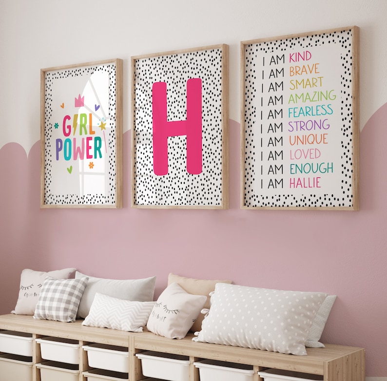 May include: Three framed art prints with a light wood frame. One print says "GIRL POWER" in colorful text. Another features a large pink letter "H". The third print lists positive affirmations. The background of the prints is white with black polka dots.